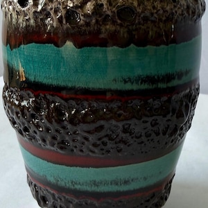 May include: A ceramic vase featuring a cylindrical shape with alternating horizontal bands of turquoise, dark brown, and deep red. The surface displays a mix of smooth, crackled glaze and thick, textured, porous brown lava-like relief sections.