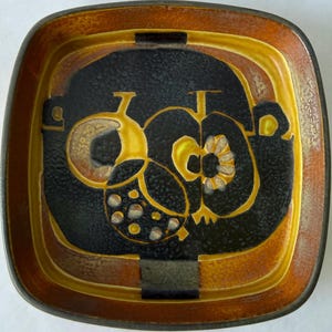 May include: A square ceramic dish featuring a dark black central area with abstract yellow and orange fruit-like motifs. The border is a gradient of warm orange and brown tones with a textured, speckled finish throughout the surface design.