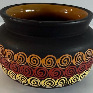 May include: A matte black ceramic bowl with a wide, flared rim and a smooth, glossy amber-colored interior. The exterior features three horizontal rows of hand-painted spiral patterns in vibrant shades of orange, deep red, and bright yellow.