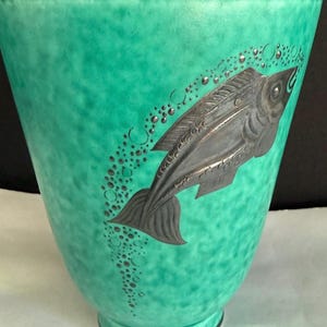 Könnte beinhalten: Eine konische Vase mit marmorierter türkiser und grüner Oberfläche. Das Äußere zeigt ein erhabenes, metallisch silberfarbenes Relief eines nach oben schwimmenden Fisches, begleitet von einer Spur kleiner, kreisförmiger Blasen zum Rand hin.