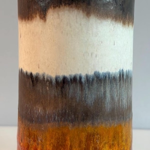 May include: A cylindrical ceramic vase featuring a multi-colored glaze with horizontal bands of dark brown, cream, grey, orange, and reddish-brown. A small rectangular sticker with the text "heuric" is adhered to the upper portion of the vase.