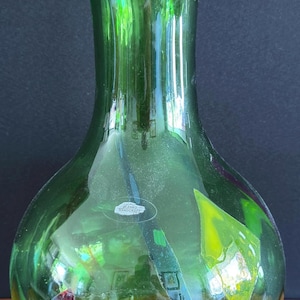 Pode incluir: Um vaso de vidro verde translúcido com borda alargada e corpo arredondado. O vaso apresenta fragmentos de vidro decorativos incrustados em amarelo e roxo escuro, e um pequeno adesivo branco com o texto Blenko Handcraft na superfície frontal.