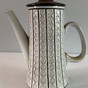 Könnte beinhalten: Eine hohe Kaffeekanne aus Keramik mit weißem zylindrischem Körper, der ein sich wiederholendes vertikales Muster aus dunkelbraunen Linien und kreisförmigen Prägungen aufweist. Sie hat einen dunkelbraunen Deckel, einen Henkel und einen Ausguss.