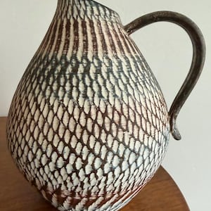 May include: A ceramic pitcher with a rounded body and a curved handle. The surface features a textured, repeating cross-hatched pattern in shades of dark brown, charcoal gray, and off-white, creating a visual effect similar to scales or woven fibers.
