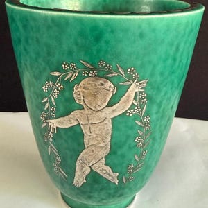 May include: A tapered ceramic vase with a mottled emerald green glaze. The exterior features a central silver-toned relief of a cherub figure framed by a delicate floral garland. The rim has a dark, thin border, and the base is circular.