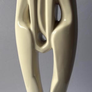 Op de afbeelding: Een hoog, crèmekleurig keramisch beeldhouwwerk dat twee abstracte figuren voorstelt die dicht bij elkaar staan. De gladde, glanzende afwerking benadrukt de langgerekte, vloeiende lijnen van de vormen die in het midden samenkomen op een ronde voet.