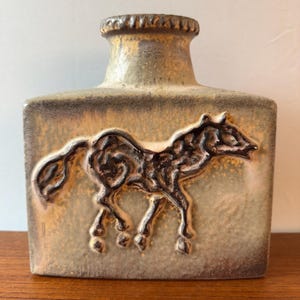 May include: A rectangular ceramic vase with a textured, mottled beige and brown glaze. The front features a raised, dark brown relief of a horse in profile. The vase has a short, cylindrical neck with a ridged rim and a flat base.
