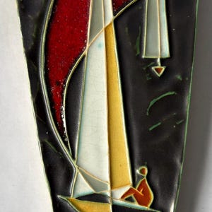 May include: A triangular wall plaque featuring a dark charcoal background with a stylized sailboat design. The boat has white and yellow sails, a red accent, and a small orange figure. The surface has a glossy finish with green edge detailing.