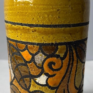 May include: A cylindrical ceramic vase with a textured mustard yellow glaze. The body features a wide horizontal band decorated with a black-outlined floral and leaf pattern in shades of burnt orange, brown, and cream, set against a yellow background.