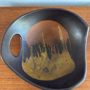 May include: A matte black ceramic bowl with an asymmetrical, organic shape and a single integrated handle. The interior features a central decorative glaze pattern in shades of burnt orange and textured mustard yellow that drips downward.