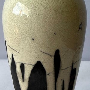Puede incluir: Un jarrón de cerámica con cuello estrecho y cuerpo bulboso que presenta una sección superior de color crema con un acabado de esmalte craquelado. La parte inferior muestra una base negra contrastante con patrones de goteo.