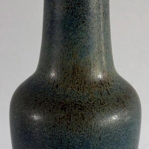 May include: A tall ceramic vase with a narrow neck and a flared rim. The body features a mottled blue and brown glaze with a textured finish. The base is decorated with a series of raised, rectangular panels, each containing a distinct geometric pattern.