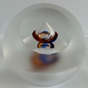 May include: A spherical glass paperweight with a frosted exterior finish. The center features a clear, pointed conical shape resting on a base of layered amber and cobalt blue glass, creating a reflective, multi-colored internal visual effect.