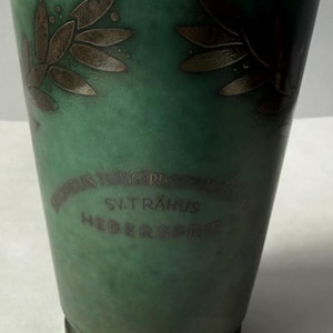 Puede incluir: Un jarrón cónico de metal verde con un motivo de hojas de laurel en metal oscuro cerca del borde. La superficie frontal muestra un texto grabado que dice SV. TRÄHUS HEDERSPRIS, con texto adicional parcialmente visible sobre la inscripción.