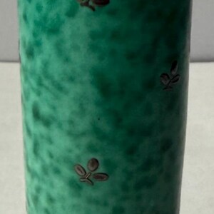 Könnte beinhalten: Eine hohe, zylindrische grüne Vase mit einer gesprenkelten Oberfläche. Das Design zeigt kleine, dunkelbraune blattförmige Motive, die über den Körper verteilt sind. Die Vase hat einen dunkelbraunen Rand oben und einen passenden Fuß.