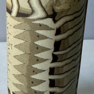 May include: A cylindrical ceramic vase featuring a beige and tan surface with dark brown, jagged, sawtooth-like patterns and horizontal lines. The exterior has a textured, matte finish with visible speckles and organic, hand-drawn geometric shapes.