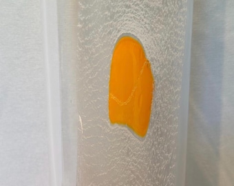 May include: A tall, cylindrical clear glass vase featuring a vertical band of frosted, textured glass. Embedded within this frosted section are four distinct, smooth, bright orange oval shapes. The base of the vase is marked with the text Kosta Boda.