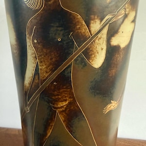 Könnte beinhalten: Eine hohe, zylindrische Keramikvase mit einer dunkelbraunen und gesprenkelten beigen Glasur. Die Oberfläche ist mit einer goldfarbenen Strichzeichnung einer Figur mit schulterlangem Haar verziert, die einen langen, dünnen Stab schräg hält.