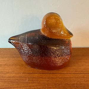 Pode incluir: Uma estatueta de pato feita de vidro cor de âmbar. O objeto apresenta uma superfície texturizada e granulada no corpo e nas asas, enquanto a cabeça e o bico são lisos e translúcidos. O vidro transita de um castanho profundo para um laranja claro.