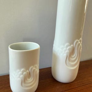 May include: A white ceramic vase and a matching smaller white ceramic tumbler. Both pieces feature a smooth, glossy finish and a raised, embossed swirling pattern around the lower section, creating a tactile, decorative texture on the surface.