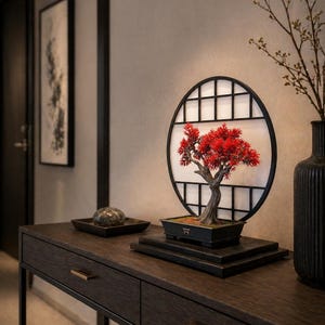 Può includere: Un bonsai decorativo con foglie rosso vivo è presentato in un vaso rettangolare nero. L'albero è esposto davanti a uno schermo a griglia rotondo bianco e nero. La scena è ambientata su una consolle in legno scuro.