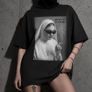 May include: Black oversized t-shirt featuring a black and white image of a nun wearing sunglasses and smoking a cigarette. The text "BERLIN 4:20 AM" is above the image. The model is wearing a black balaclava and sunglasses.