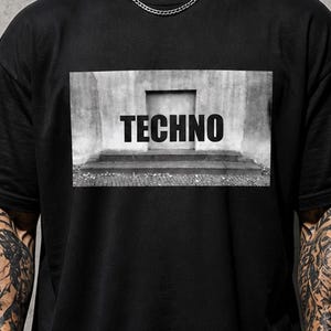 Può includere: T-shirt nera con grafica in scala di grigi con la parola "TECHNO" in grassetto. La grafica raffigura una struttura in cemento con gradini. La maglietta ha maniche corte e girocollo.