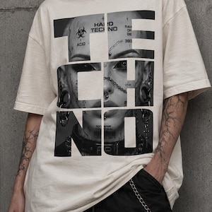 May include: An off-white short-sleeve t-shirt featuring a large black and white graphic print of a face with facial piercings and tattoos. The word TECHNO is printed in large bold letters over the image, which includes text like ACID, HARD TECHNO, and RAVER OR DIE.