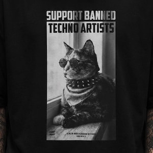 May include: Black t-shirt featuring a black and white image of a cat wearing sunglasses, a studded collar, and a bandana. The text "SUPPORT BANNED TECHNO ARTISTS" is printed above the cat. Below the cat are additional text elements.