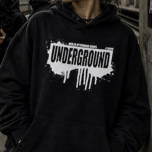 Peut inclure: Un sweat à capuche noir avec un graphisme blanc « UNDERGROUND » en lettres stylisées. Le texte est placé sur un fond blanc éclaboussé. Le sweat à capuche a une capuche à cordon et une poche avant. Le texte « BERLIN AFTERHOUR SERIES // 003AM » est également visible.