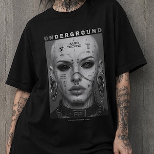 May include: A black short-sleeved t-shirt featuring a large rectangular graphic print on the chest. The print displays a grayscale portrait of a person with facial piercings, tattoos, and text including "UNDERGROUND," "HARD TECHNO," and "RAVE OR DIE."