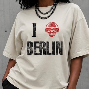 May include: Beige t-shirt with the text "I Berlin" in black, featuring a red skull graphic. The shirt has short sleeves and a relaxed fit. The model is wearing a silver chain necklace.