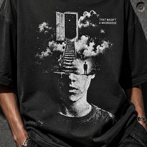 Op de afbeelding: Een zwart T-shirt met korte mouwen en een witte grafische print van een menselijk hoofd met een trap die naar een open deur in de wolken leidt. De tekst "THAT WASN'T A MICRODOSE" is in het wit gedrukt in de rechterbovenhoek van de afbeelding.