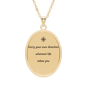 Peut inclure: Un collier pendentif ovale doré avec une petite icône de rose des vents noire en haut et le texte noir gravé "Carry your own direction wherever life takes you" centré sur la surface lisse et mate du pendentif.