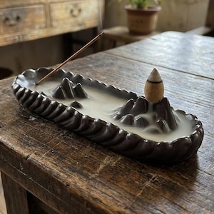 Puede incluir: Un porta incienso de cerámica marrón oscuro con un diseño de cordillera. El humo del incienso se eleva del soporte, y un palito de incienso encendido se coloca en el soporte. Un cono de incienso de madera se encuentra en la cima de la cordillera.