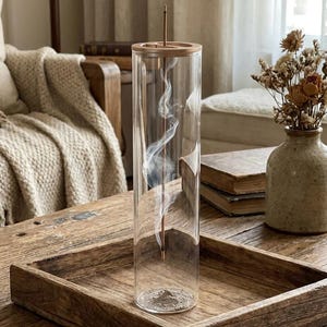 Könnte beinhalten: Ein klarer Glas-Räucherstäbchenhalter mit Holzaufsatz und einem brennenden Räucherstäbchen, das weißen Rauch abgibt. Der Halter steht auf einem Holztablett. Rechts steht eine Vase mit Trockenblumen.