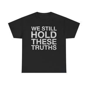 Puede incluir: Camiseta negra con el texto blanco "WE STILL HOLD THESE TRUTHS" en una fuente audaz y en bloques. La camiseta es de estilo cuello redondo clásico, adecuada para uso casual. El texto está centrado en el pecho.