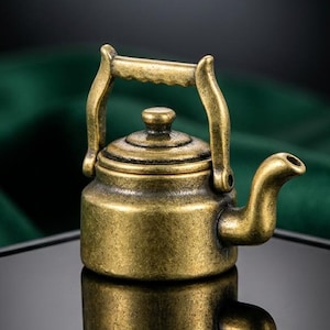 Op de afbeelding: Een kleine, antieke messing theepot met een gebogen tuit, handvat en deksel. De theepot staat op een reflecterend oppervlak, met een donkergroene stof op de achtergrond. Het ontwerp suggereert een vintage of verzamelobject.
