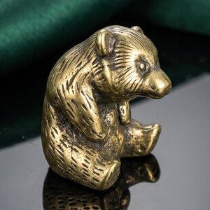 May include: A small, brass-colored bear figurine in a seated position. The bear has detailed fur texture and is sitting with its paws clasped. The figurine is on a reflective surface with a dark green backdrop.