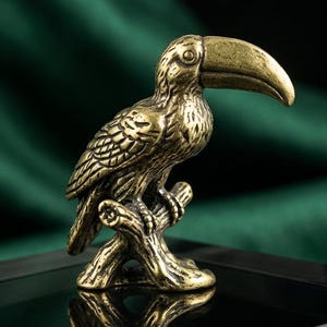 May include: A detailed brass toucan figurine perched on a branch. The bird has a large, curved beak and textured feathers. The figurine is displayed on a black surface against a dark green backdrop.