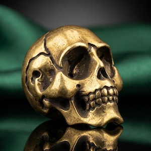 Puede incluir: Una figura de calavera dorada con detalles intrincados y grietas visibles. La calavera se refleja en la superficie. El fondo es una tela verde oscuro drapeada, creando un contraste con el objeto.