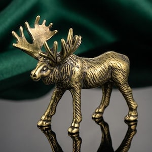 May include: A detailed bronze-colored moose figurine with large antlers stands on a reflective black surface. The moose is posed in a walking stance, with intricate detailing on its body. The background features a dark green, textured fabric.