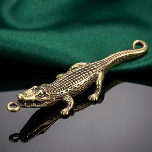 May include: A detailed brass alligator figurine with a textured body and a small loop at the snout. The reptile is posed on a reflective surface, with a dark green fabric in the background.