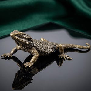 Peut inclure: Une figurine détaillée de lézard barbu de couleur bronze. Le reptile est représenté avec ses pattes tendues, semblant ramper. La figurine est posée sur une surface noire réfléchissante, avec un tissu vert foncé en arrière-plan.