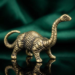 Könnte beinhalten: Kleine Dinosaurierfigur in Messingoptik mit langem Hals und Schwanz. Die detaillierte Skulptur weist eine strukturierte Haut auf und ist vor einem dunkelgrünen Hintergrund platziert. Der Dinosaurier ist etwa 10 cm lang.