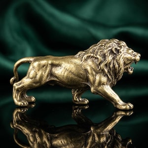 Op de afbeelding: Een gedetailleerd bronzen leeuwenbeeldje in een loopstand, met de mond open in een brul. De sculptuur heeft een getextureerde manen en lichaam, die licht reflecteren. Het beeldje staat tegen een donkere, reflecterende ondergrond en een groene stoffen achtergrond.