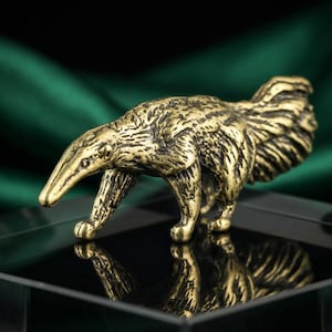May include: A detailed, antique-style brass anteater figurine. The sculpture is posed in a walking stance, with intricate detailing on its fur and body. The figurine is displayed on a black surface against a dark green backdrop.