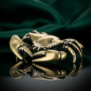 May include: A detailed brass crab figurine with a textured surface. The crab has large claws and multiple legs, with a reflective surface beneath it. The background is a dark green fabric.
