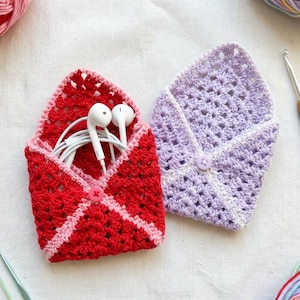 Puede incluir: Dos pequeñas fundas de ganchillo en forma de sobre, una de color rojo vibrante con borde rosa y la otra de color morado claro. Cada funda tiene un cierre de botón en la solapa central. Unos auriculares blancos con cable están dentro.