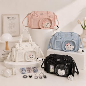 Op de afbeelding: Vier handtassen in roze, wit, blauw en zwart, elk met een strik en een doorzichtig venster met een pluche puppy. De tassen hebben zilverkleurige hardware en omvatten een tote, een tas en een crossbody-stijl. Accessoires zijn sleutelhangers en pins.